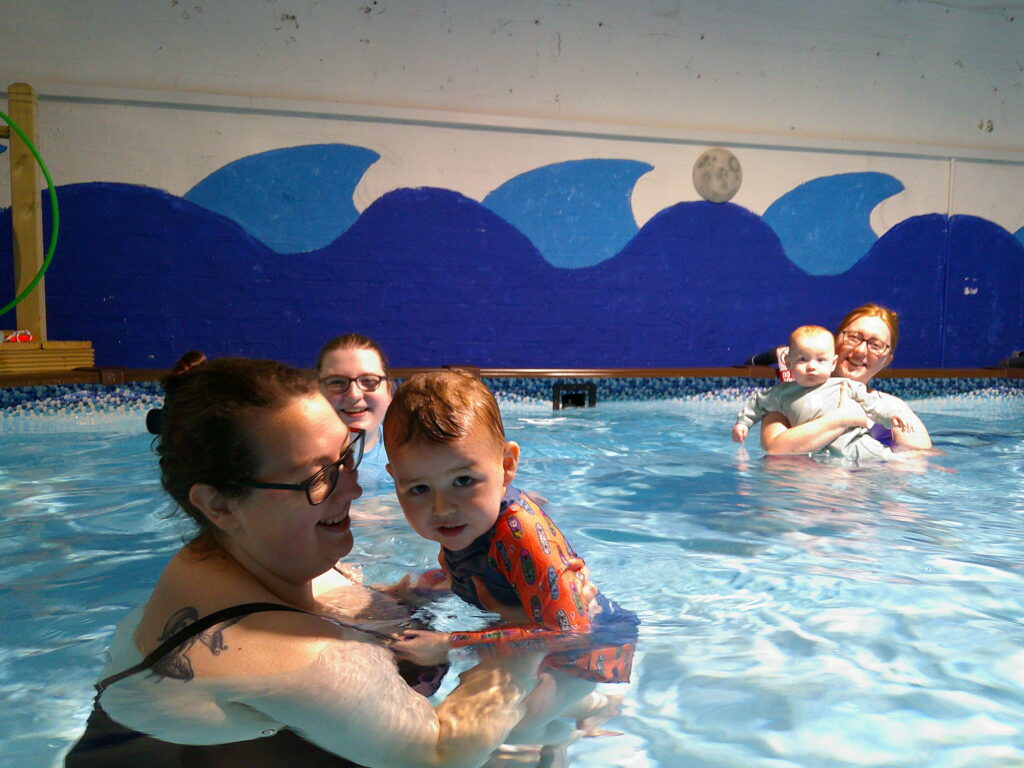 Adults and young children are enjoying a swim in an indoor pool with a blue wave mural on the wall behind them. One woman is holding a toddler in the foreground, while another holds a baby in the background.