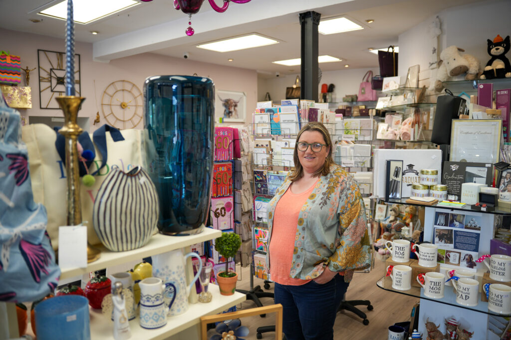 A woman wearing glasses and a floral kimono stands smiling in a colorful gift shop filled with mugs, vases, books, and various decorative items arranged on shelves and tables.