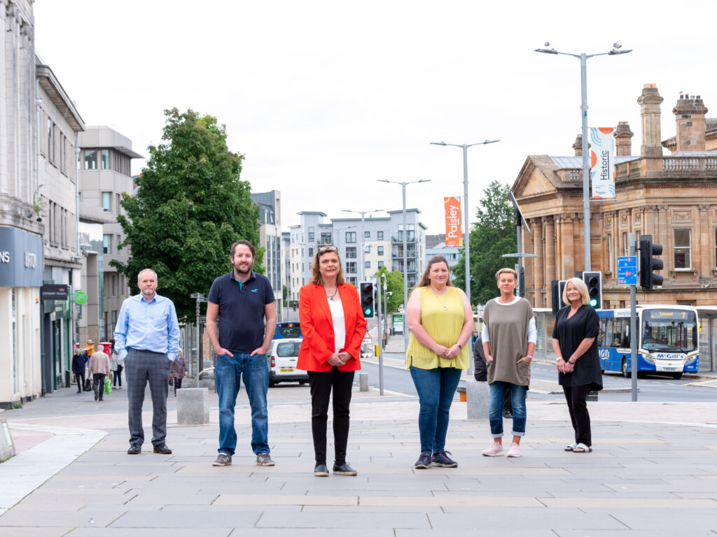 Six people stand spaced apart in a pedestrian city street with buildings, traffic signs, and a bus in the background; the weather is overcast and the group faces the camera.