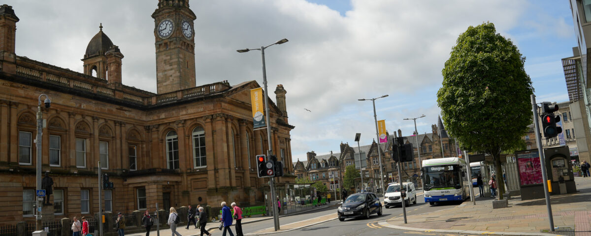 Pedestrians cross a street at a crosswalk near a historic building with clock towers. A bus waits nearby and traffic lights are visible. The sky is partly cloudy and trees line the street.