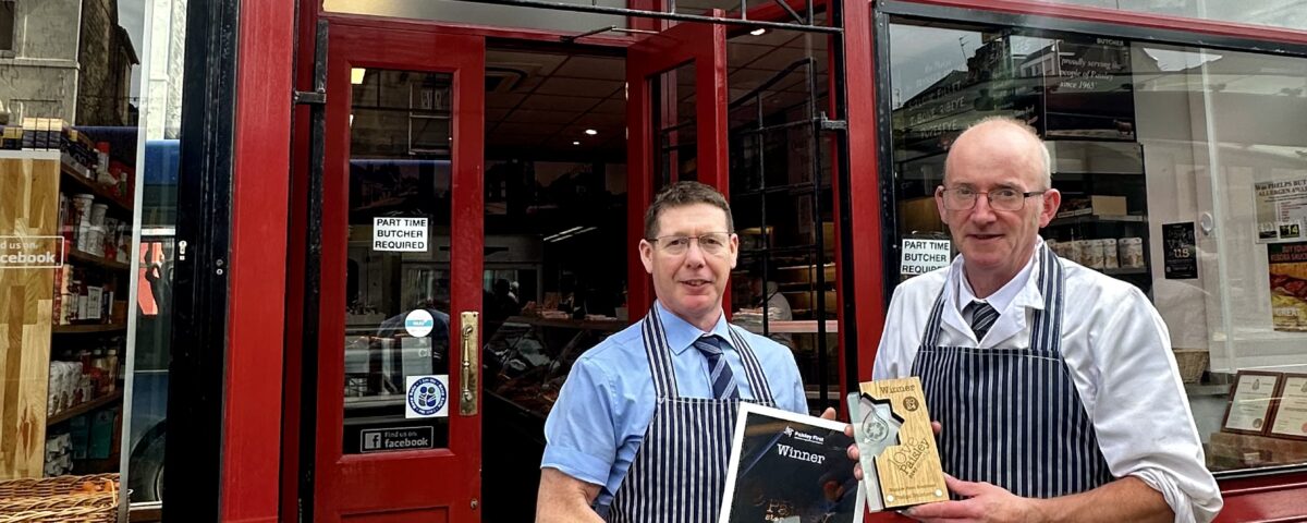Two butchers in striped aprons stand outside WM. Phelps butcher shop, smiling and holding framed awards. The shop has a red door, tiled entrance, and special offer signs displayed.