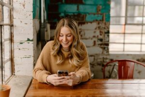 A young woman with long blonde hair, wearing a tan sweater, sits at a wooden table by a window and smiles while looking at her smartphone in a rustic, brightly lit cafe.