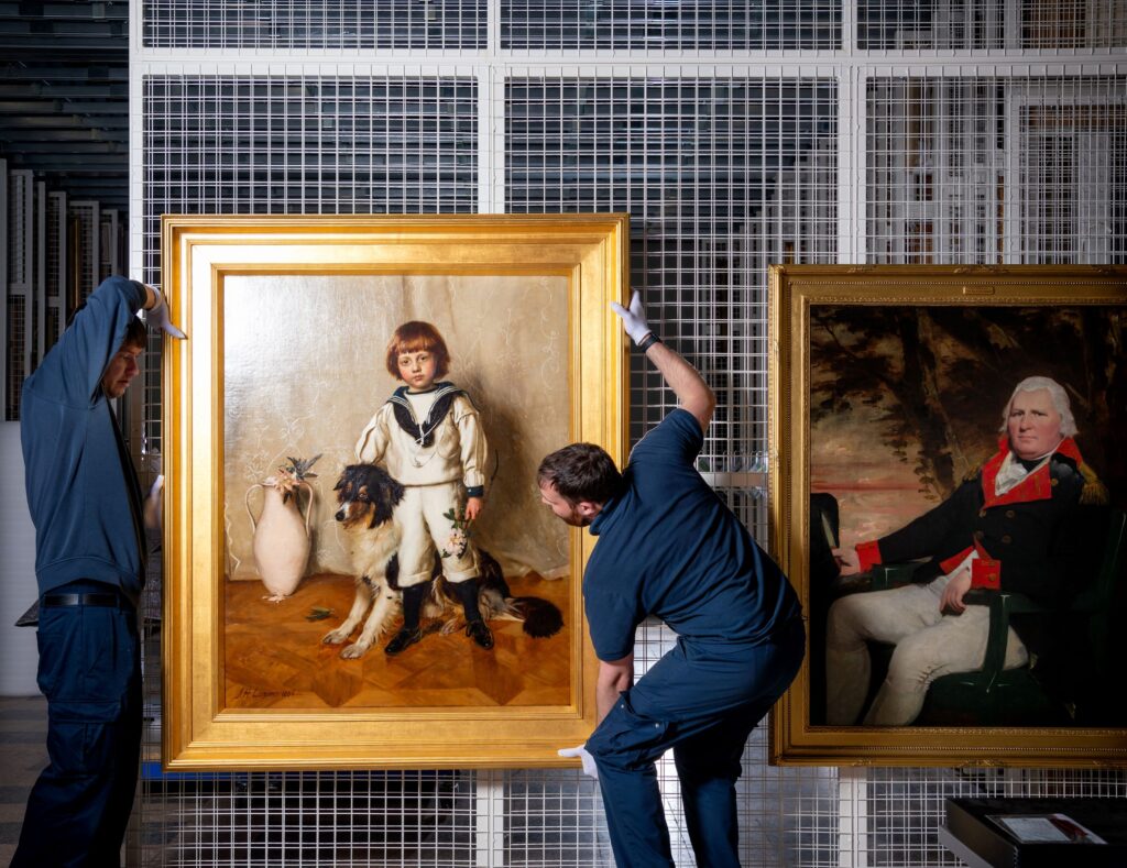 Two workers in blue uniforms carefully hang a large framed painting of a child with a dog, next to another portrait of a seated man in uniform, in an art storage area with metal grid walls.