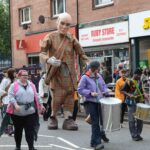 A street parade features a giant human puppet in plaid clothing, followed by people playing drums. Bystanders watch beside shops, including Ruby Store and Subway, on a city street.