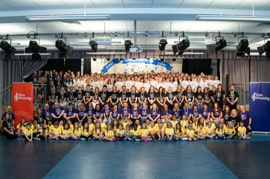 A large group photo of children and adults arranged in rows on a stage and floor, all wearing colorful t-shirts in yellow, purple, black, and white. Two banners on each side read “Feis Phàislig”. The background shows a blue and white sign.
