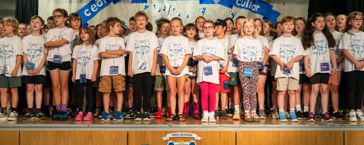 A group of children stand on a stage singing together, wearing matching white T-shirts. Behind them is a banner with “ceilidh,” “coimhearsnachd,” and “cultural.” The school’s logo is visible on the front of the stage.