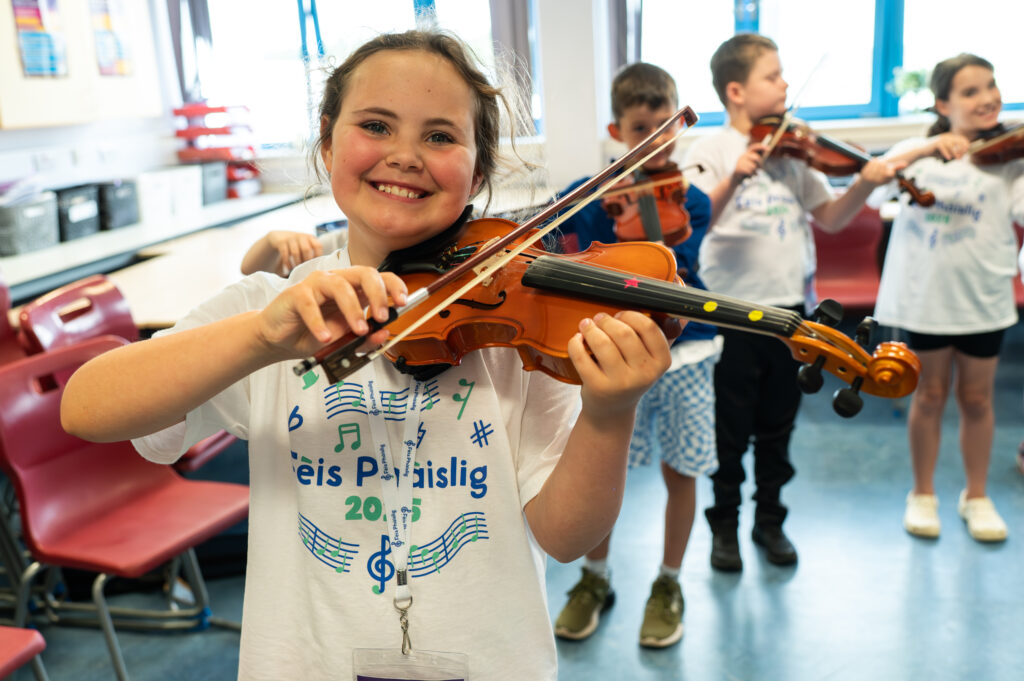 A smiling girl plays the violin in a classroom, wearing a white T-shirt with a music-themed design. Behind her, three other children also play violins, all appearing happy and engaged in the music activity.