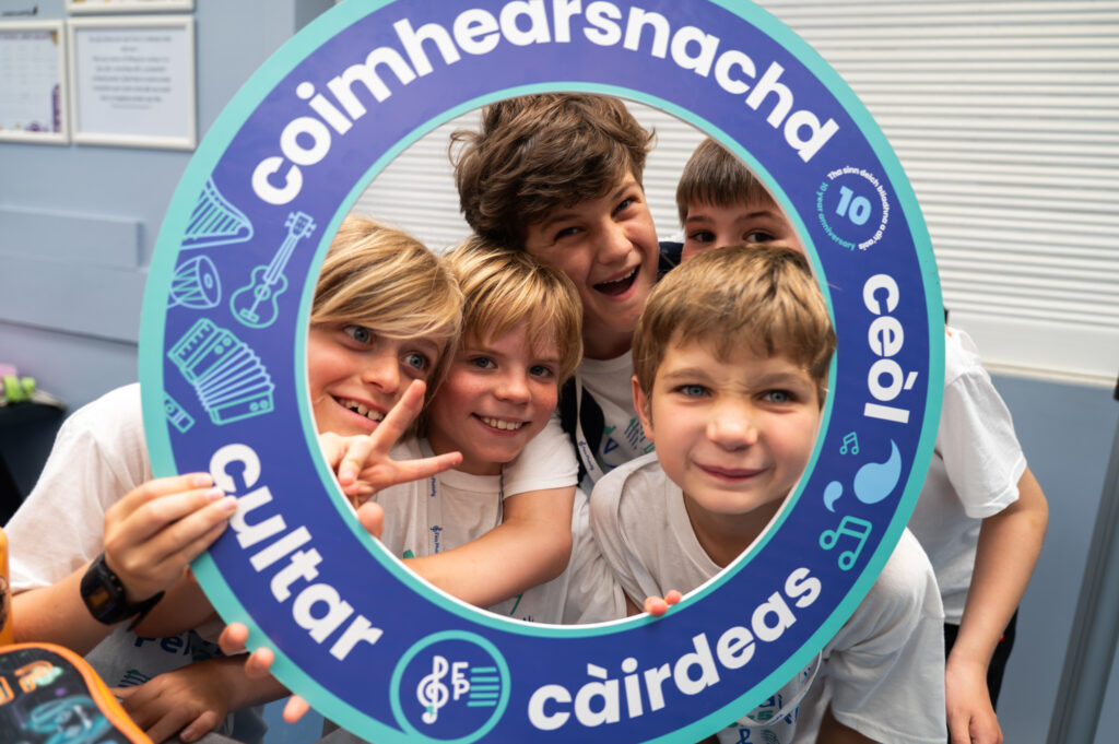 Five smiling boys pose together, looking through a large blue circular frame with Gaelic words and music symbols. The boys wear white shirts and appear cheerful and excited in a bright indoor setting.