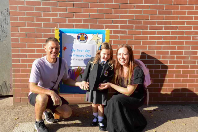 A smiling young girl in a school uniform stands between two adults in front of a brick wall and a colorful sign that reads “My first day in Primary One.” The adults are crouching and holding the girl's hands.