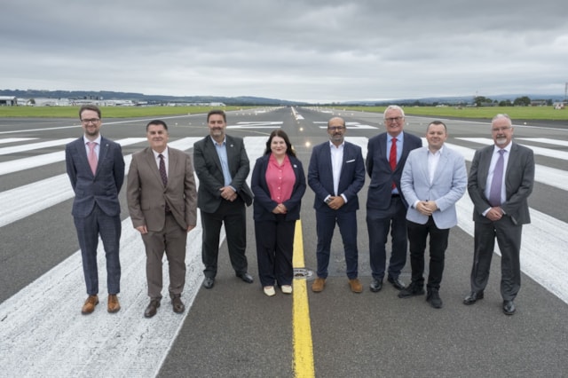 Nine people in business attire stand side by side on the center line of an airport runway under a cloudy sky, with green fields and distant buildings in the background.