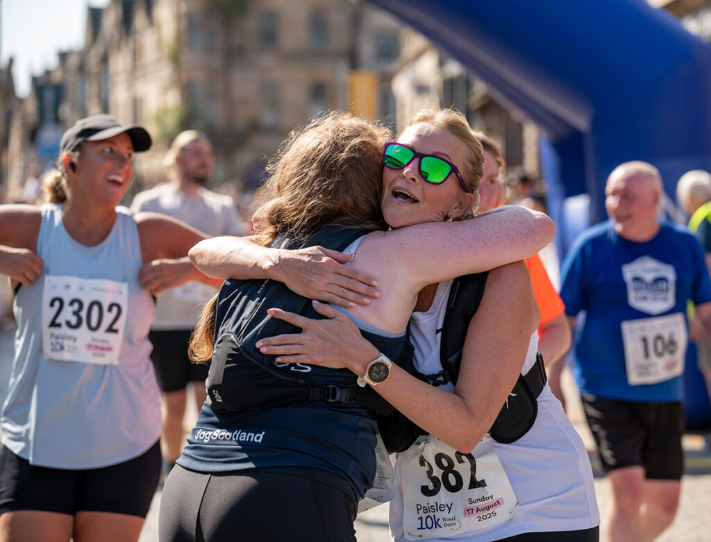 Two women, both wearing running gear and race numbers, embrace joyfully at the finish line of a race. Other runners with race numbers smile and walk in the background on a sunny day.