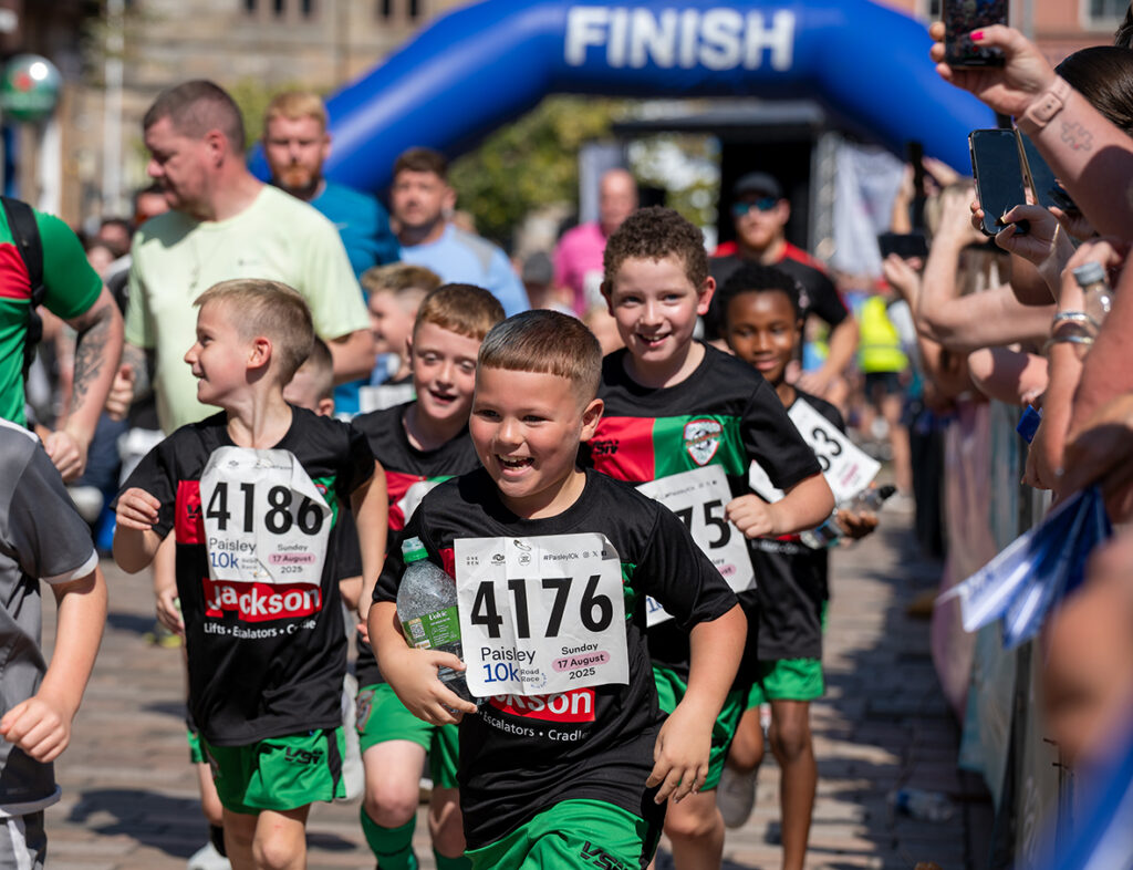 A group of smiling children in sportswear run toward the finish line at a race event, with spectators cheering on both sides and a blue "FINISH" arch visible in the background.