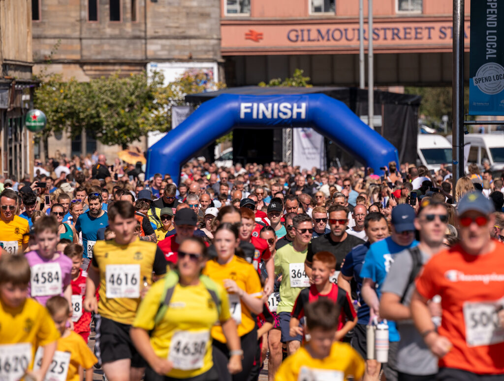 A large crowd of runners, many wearing numbered bibs and colorful shirts, approaches a blue "FINISH" archway during a city race event on a sunny day.