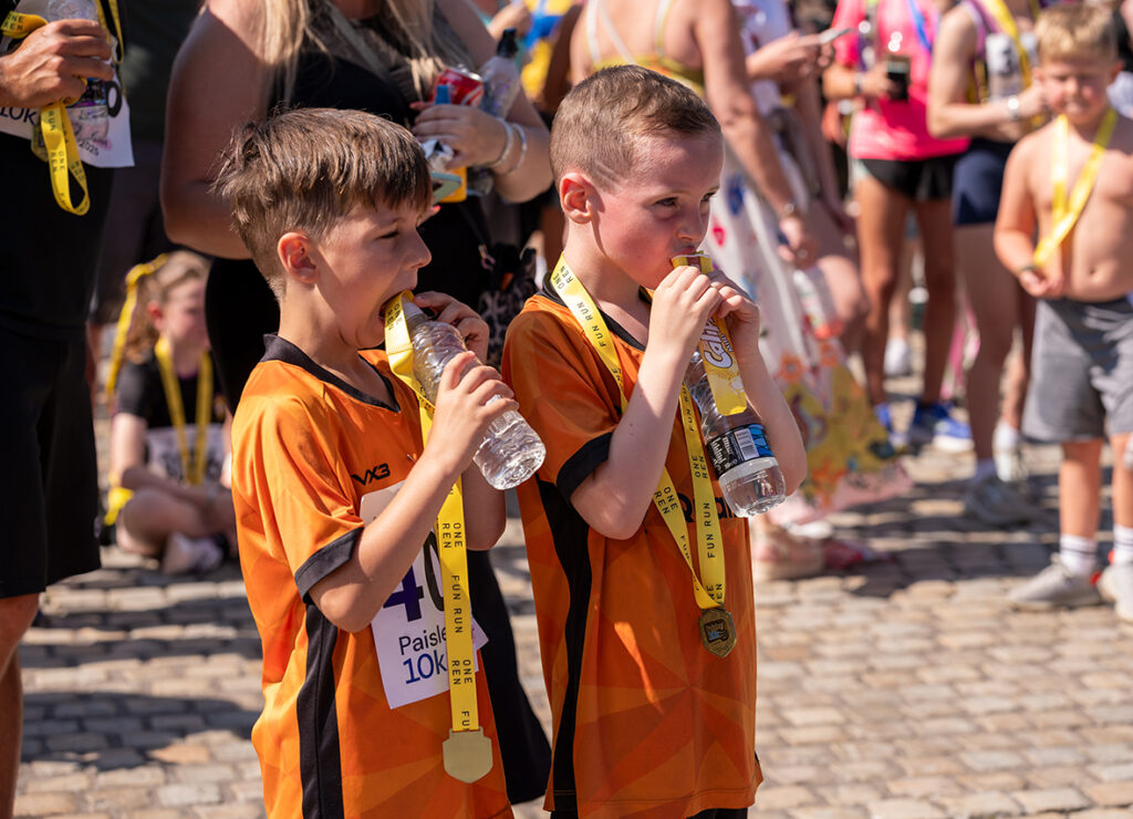 Two young boys in orange shirts stand side by side at a race event, wearing medals and drinking water. A crowd of people with medals and race numbers gathers in the sunny background.
