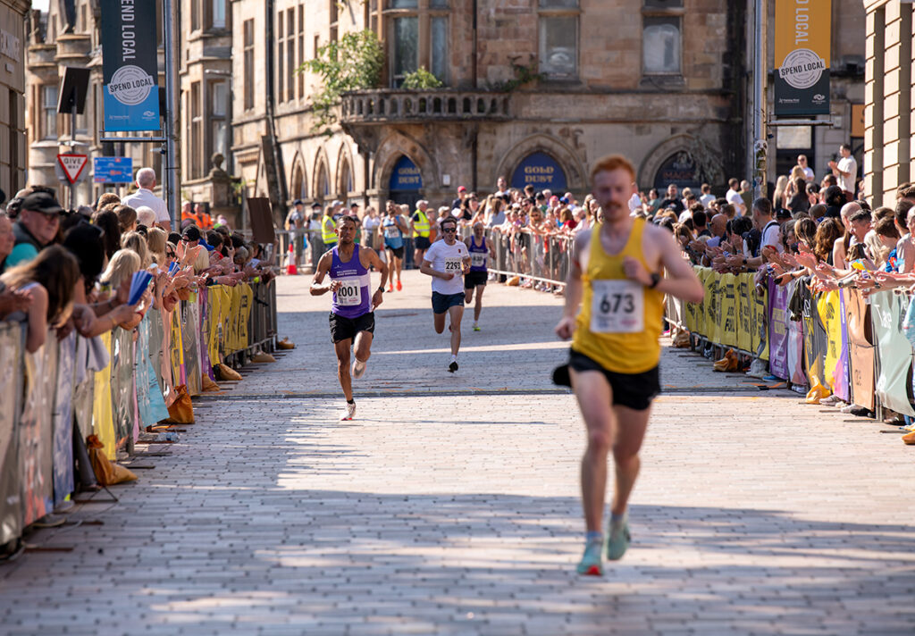 Runners race down a sunlit city street lined with cheering crowds behind barriers. One runner in a yellow shirt leads, with three others trailing behind. Historic buildings and banners are visible in the background.