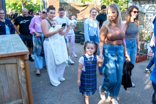 A smiling woman holding a girl’s hand leads a group of adults and children outdoors. The girl wears a blue plaid school uniform, while others walk behind, some carrying babies and chatting, on a sunny day.