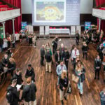 A large group of people gathers in a spacious hall with wooden floors, red curtains, and exhibition tables. A screen at the front displays a SpongeBob meme and event information.