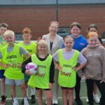 A group of smiling children and a woman stand in front of a football goal on an outdoor sports field. Most of the kids wear yellow sports bibs, and one girl holds a purple and white soccer ball.