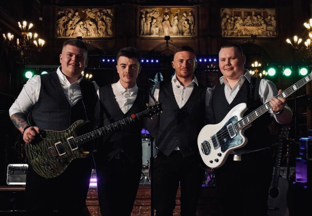 Four men dressed in white shirts, dark vests, and trousers stand together on stage, two holding electric guitars, with instruments and ornate wood carvings visible in the background.