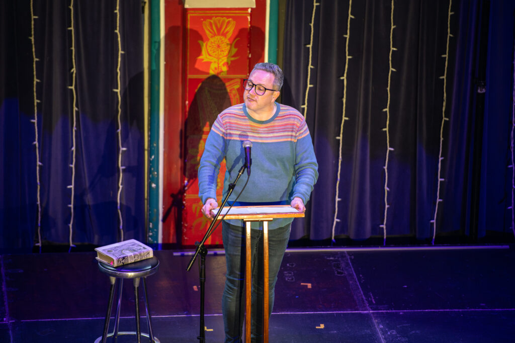 A person wearing glasses and a striped sweater stands at a podium with a microphone, reading or speaking. A closed book rests on a stool nearby. Curtains and string lights hang in the background.