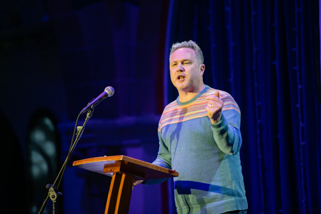 A man in a blue sweater speaks passionately at a podium on stage, with a microphone in front of him and blue curtains in the background.