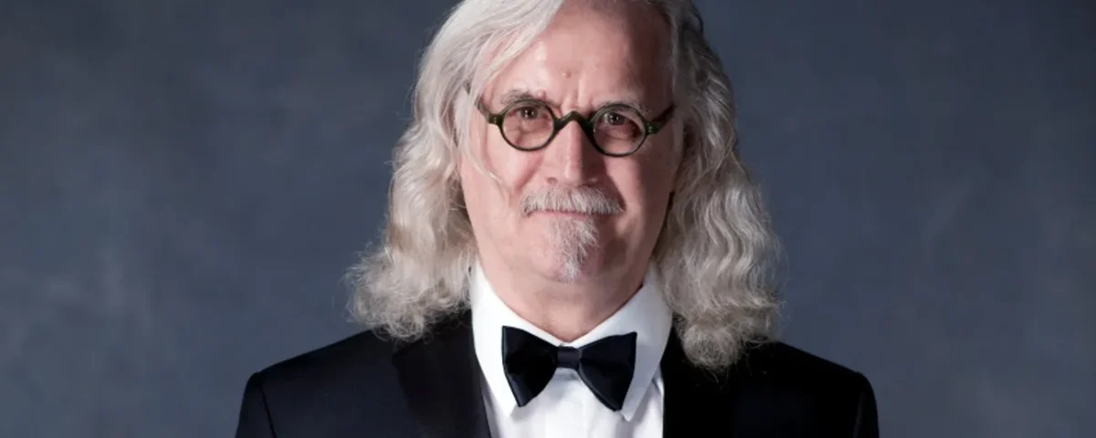 An older man with long, wavy gray hair and glasses, wearing a black tuxedo with a bow tie, stands in front of a dark gray background, smiling slightly.