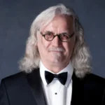 An older man with long, wavy gray hair and glasses, wearing a black tuxedo with a bow tie, stands in front of a dark gray background, smiling slightly.