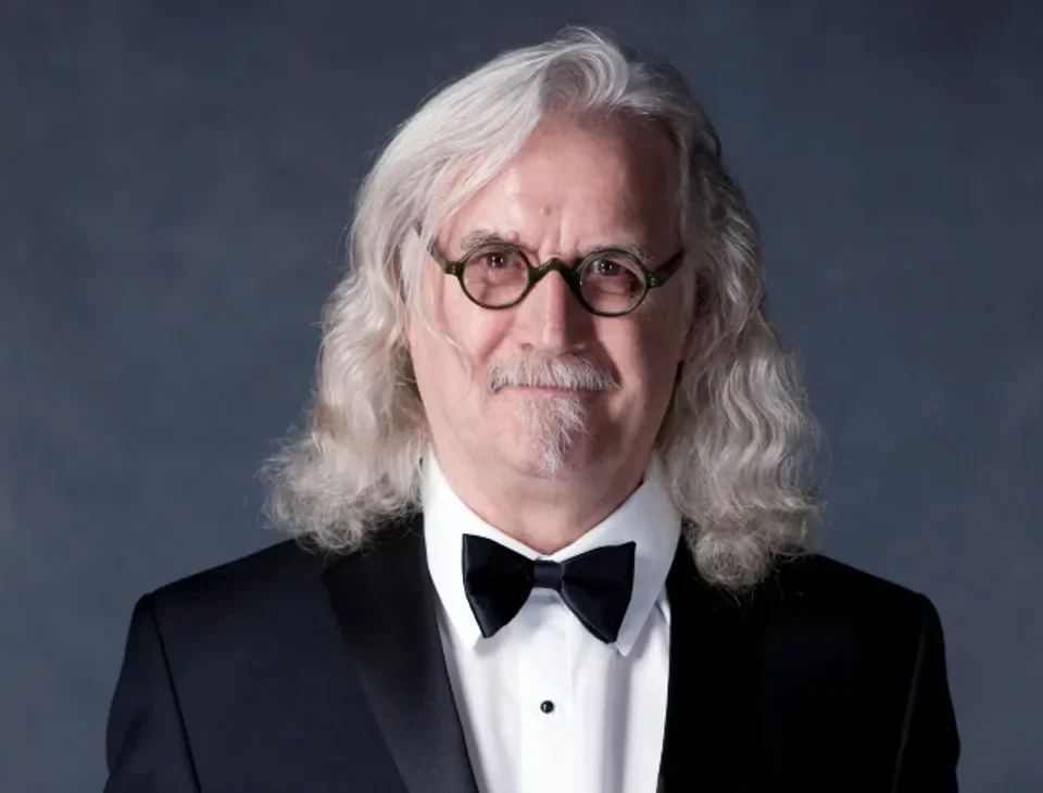 An older man with long, wavy gray hair and glasses, wearing a black tuxedo with a bow tie, stands in front of a dark gray background, smiling slightly.