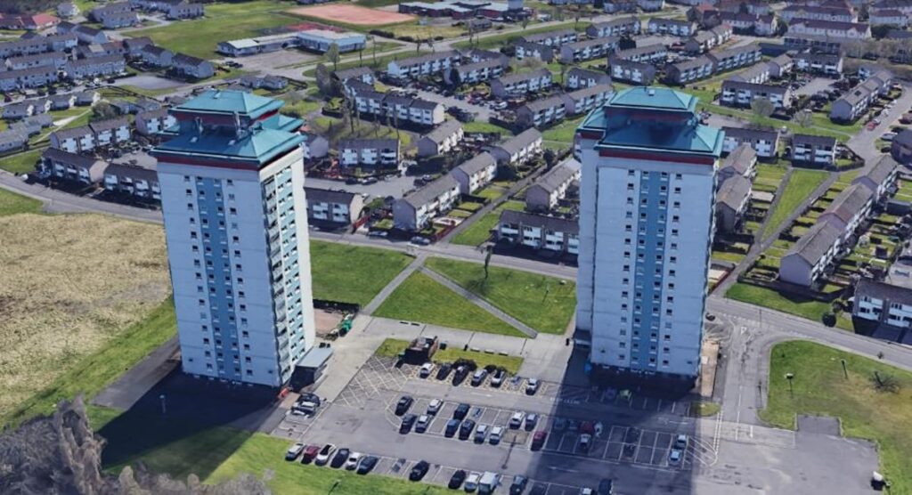 Aerial view of two tall, identical apartment towers with blue roofs, surrounded by parking lots and low-rise houses in a suburban neighborhood.
