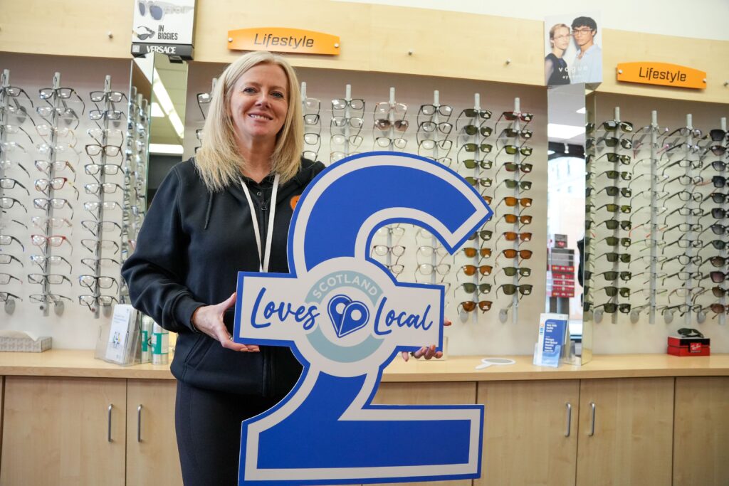 A woman stands in front of a display of eyeglasses, smiling and holding a large blue "Scotland Loves Local" sign shaped like a pound symbol inside an eyewear store.