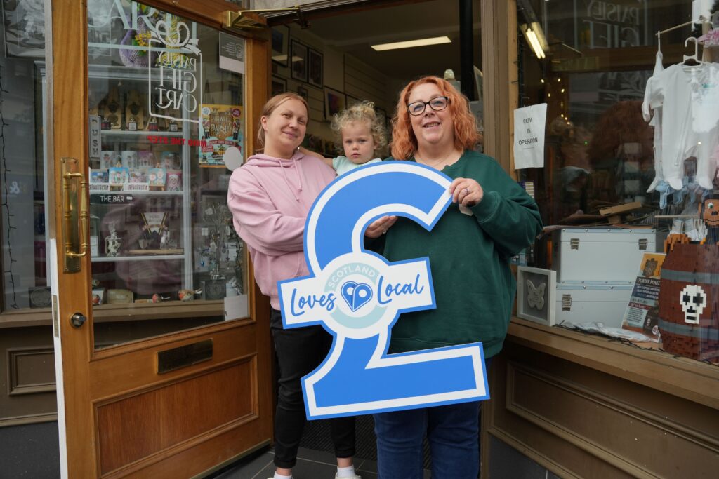 Two women and a child stand outside a shop entrance, smiling and holding a large blue pound (£) sign that says “Scotland Loves Local.” The shop window displays various items and crafts.