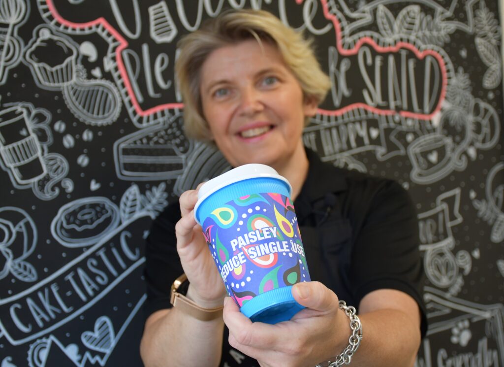 A smiling woman holds a colorful coffee cup labeled “PAISLEY PRODUCE SINGLE JUSE” in front of a blackboard decorated with doodles of desserts and cafe-themed drawings.