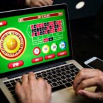 A person plays an online roulette game on a laptop, with colorful betting options and a virtual roulette wheel visible on the screen. Hands are on the keyboard, and a smartphone and other items are nearby.