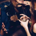 Three people are sitting close together at a party or club, laughing and talking. Two of them are holding drinks with straws and ice, and their faces show joy and engagement.
