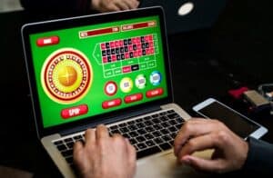 A person plays an online roulette game on a laptop, with colorful betting options and a virtual roulette wheel visible on the screen. Hands are on the keyboard, and a smartphone and other items are nearby.