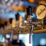 Close-up of shiny beer taps in a bar, with a blurred background of glasses and warm lighting. One tap displays a round label with the word "Supermacy.
