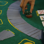 A hand reaches for a stack of poker chips on a green casino table with playing cards, face-down cards arranged in a curve, and two face-up cards: the queen of clubs and seven of hearts.