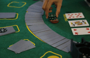 A hand reaches for a stack of poker chips on a green casino table with playing cards, face-down cards arranged in a curve, and two face-up cards: the queen of clubs and seven of hearts.