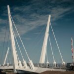 A modern white cable-stayed pedestrian bridge with tall angled supports under a blue sky. Several people are walking on the bridge, and a barrier gate is visible on the right.