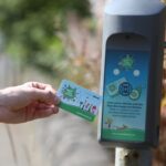 A person holds a transit card up to a card reader mounted on a pole outdoors, preparing to scan it. The card and reader display green graphics related to public transport.