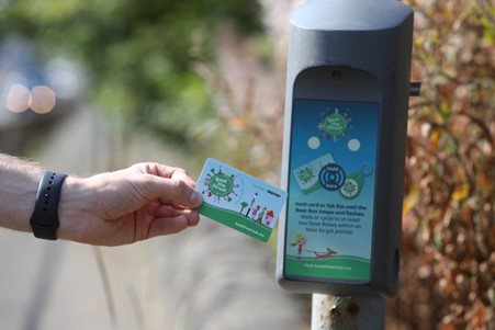 A person holds a transit card up to a card reader mounted on a pole outdoors, preparing to scan it. The card and reader display green graphics related to public transport.