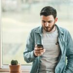 A man with a beard wearing a denim jacket over a gray shirt is leaning against a windowsill, looking at his smartphone. A small potted cactus sits on the windowsill beside him.