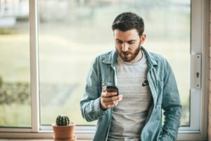 A man with a beard wearing a denim jacket over a gray shirt is leaning against a windowsill, looking at his smartphone. A small potted cactus sits on the windowsill beside him.