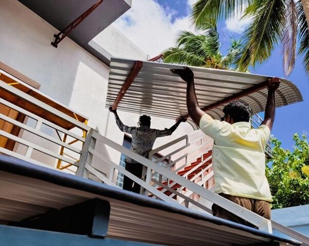 Two men lift a corrugated metal sheet onto a roof, with palm trees and blue sky in the background. One man is standing on a lower level, assisting with the placement.