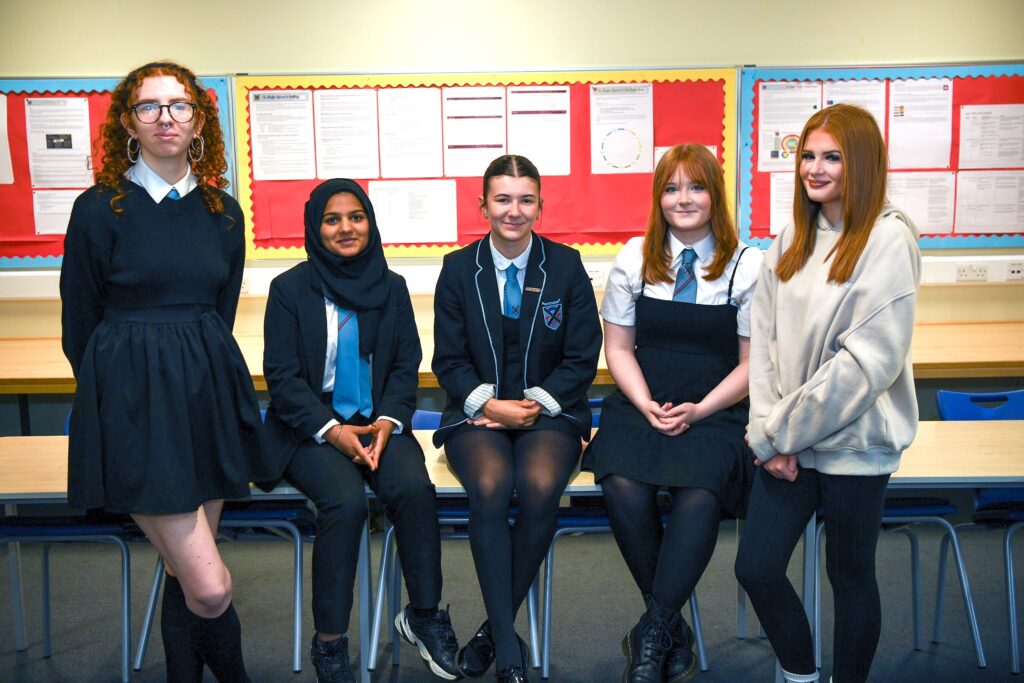 Five teenage girls are posing and smiling in a classroom, dressed in a mix of school uniforms and casual clothes, with bulletin boards and papers visible on the wall behind them.