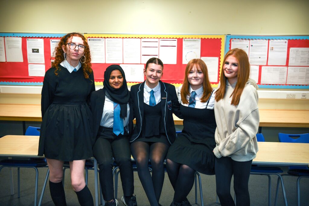 Five teenage girls pose together in a classroom, smiling. Four wear school uniforms with black and blue accents, while one wears a beige hoodie and black leggings. They stand and sit in front of a red bulletin board with papers.