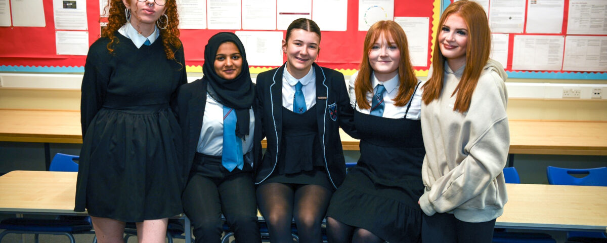 Five teenage girls, wearing school uniforms, stand and sit together in a classroom in front of red bulletin boards, smiling at the camera.