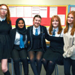 Five teenage girls, wearing school uniforms, stand and sit together in a classroom in front of red bulletin boards, smiling at the camera.