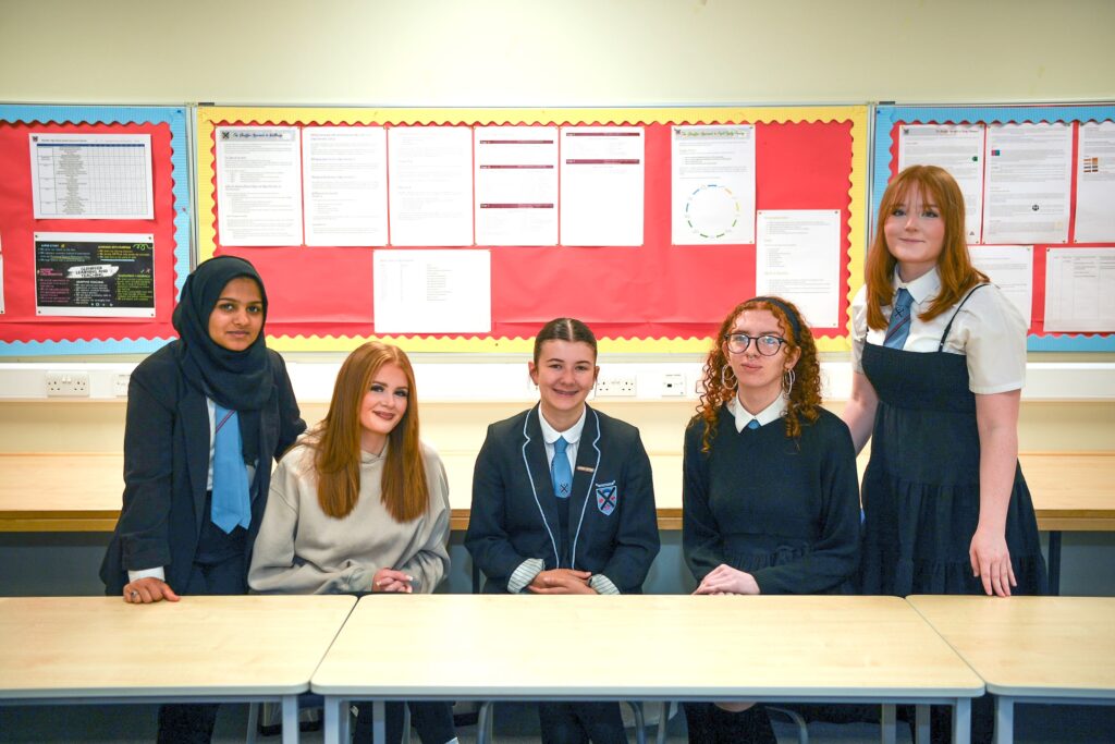 Five students sit and stand around a classroom table, facing the camera. They are dressed in school uniforms and casual clothes, with a red bulletin board displaying papers behind them.
