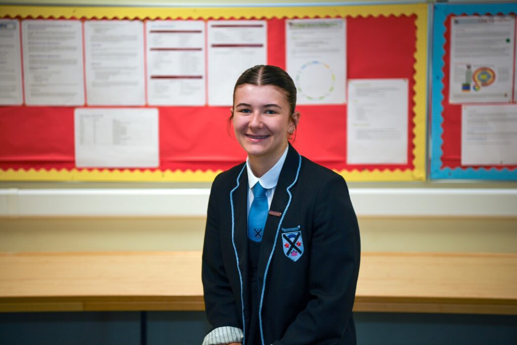 A student in a dark school blazer, white shirt, and blue tie sits and smiles in a classroom with colorful notice boards and documents displayed on the wall behind her.